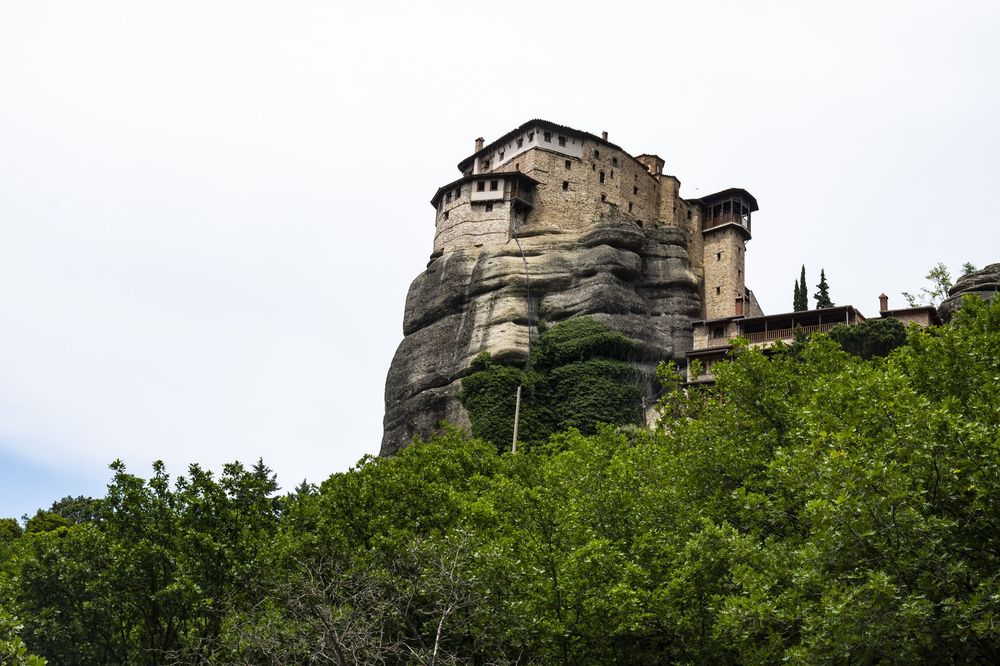 Ancient stone castle tower rises above lush green trees on a hilltop against cloudy sky.