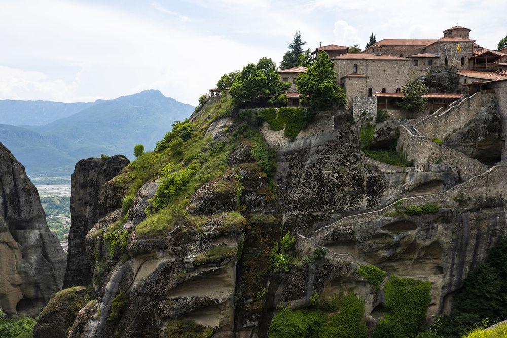 Ancient stone monasteries perched atop dramatic rocky cliffs at Meteora in Greece against a cloudy mountain backdrop.