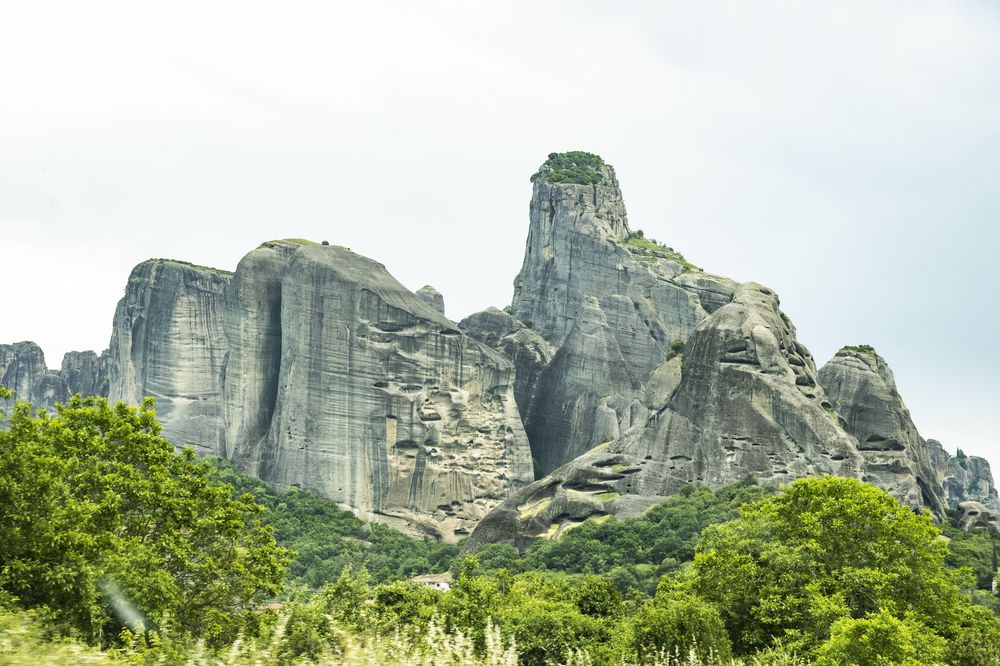 Dramatic rock formations of Meteora rise above lush green forest in Greece against cloudy sky.