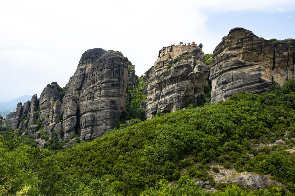 Dramatic rock formations of Meteora rise above lush green forest in Greece against a cloudy sky.