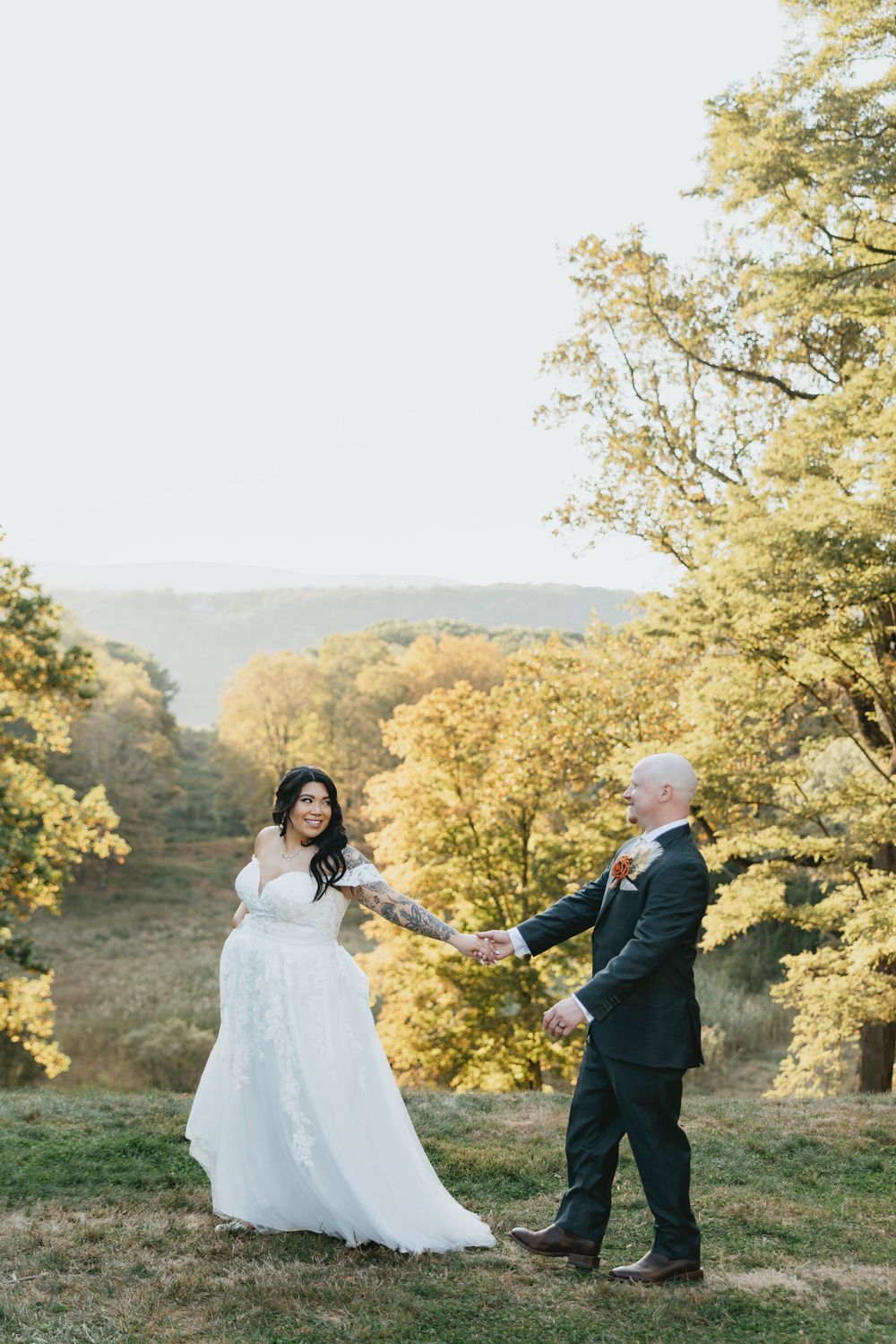 A couple in wedding attire spins and dances together on a hilltop during golden hour with trees in the background.