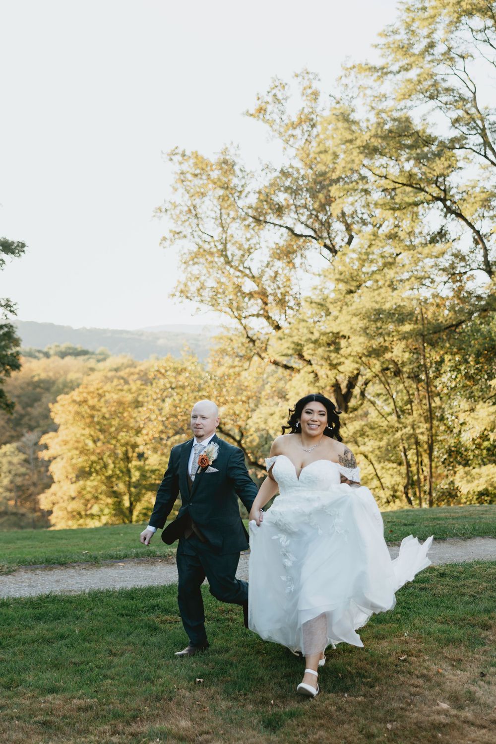 A couple runs playfully through a grassy hilltop with autumn foliage and scenic views behind them.