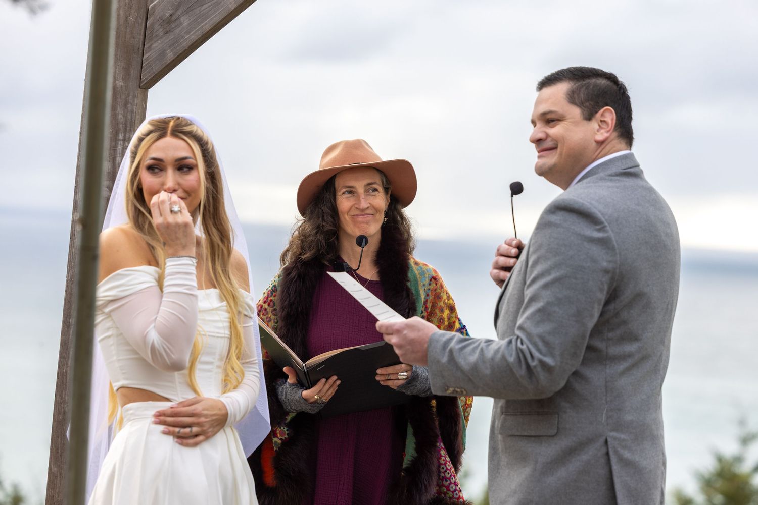 An outdoor wedding ceremony takes place with an officiant in burgundy and a couple against a cloudy sky backdrop.