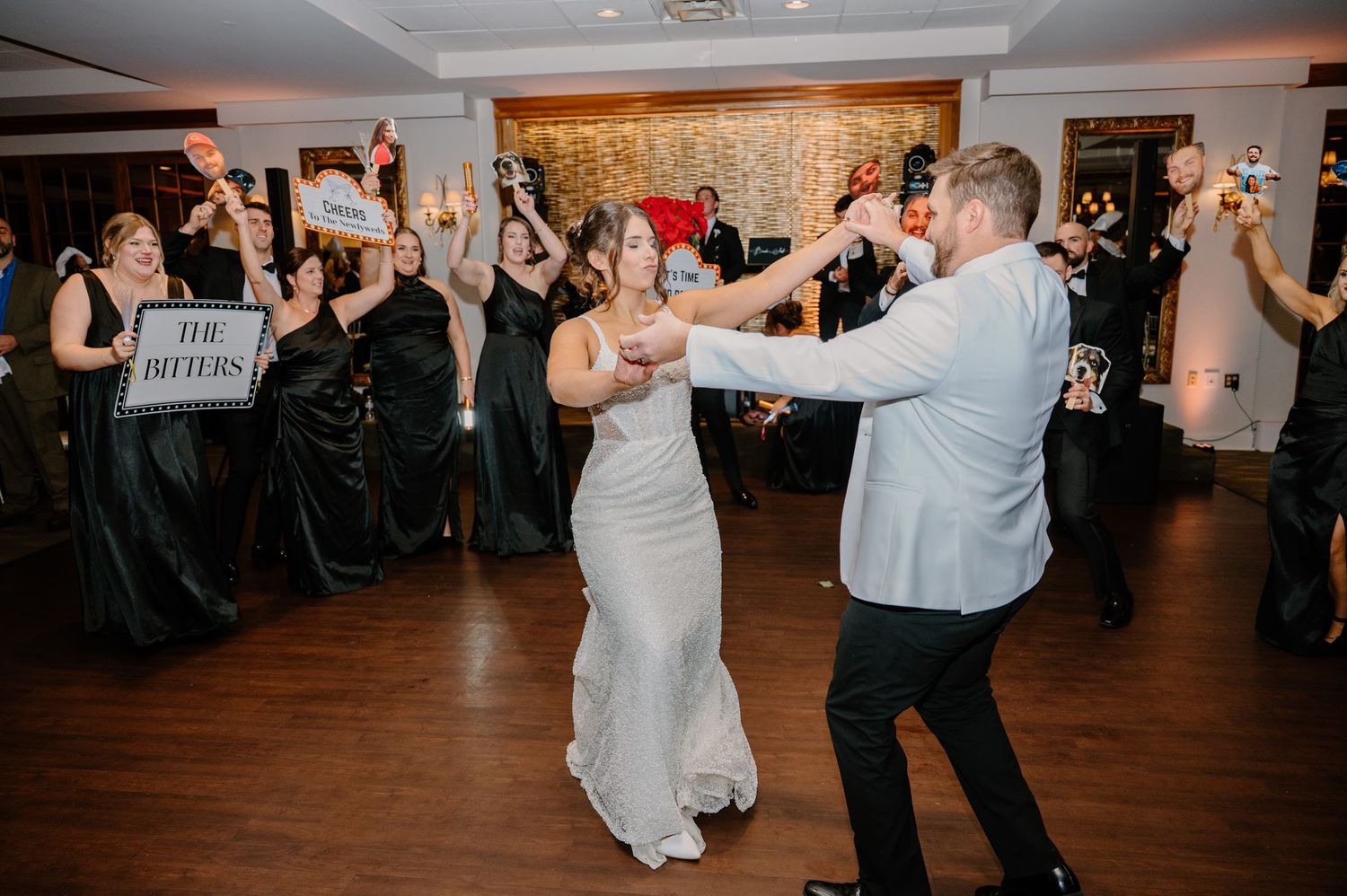 Newlyweds share their first dance on a wooden floor while wedding guests cheer and take photos in the background.