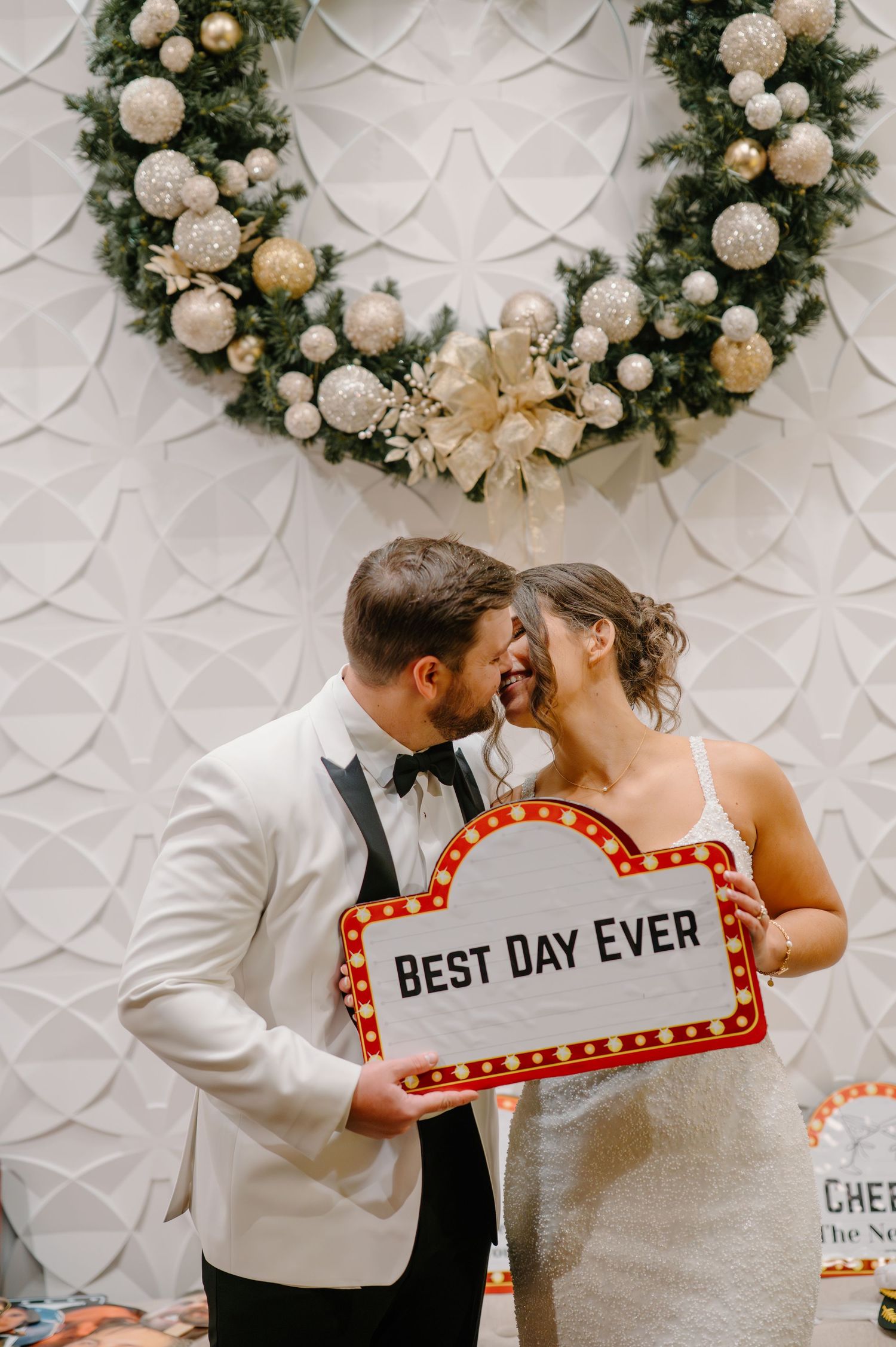 Wedding couple shares romantic moment in front of white textured wall with decorative wreath and illuminated marquee sign.