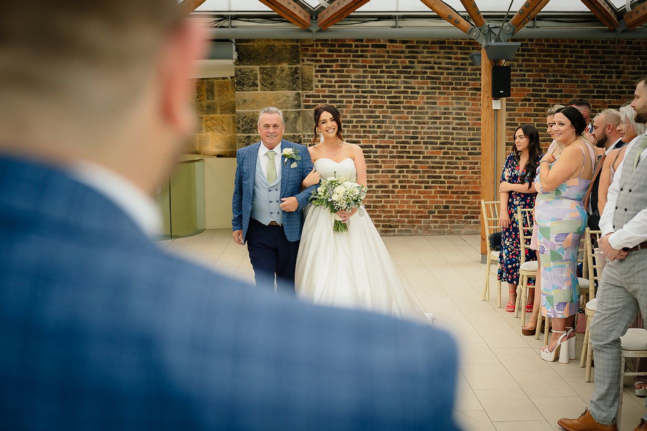 Wedding guests celebrate and dance together on a rustic wooden floor at an indoor brick-walled reception venue.