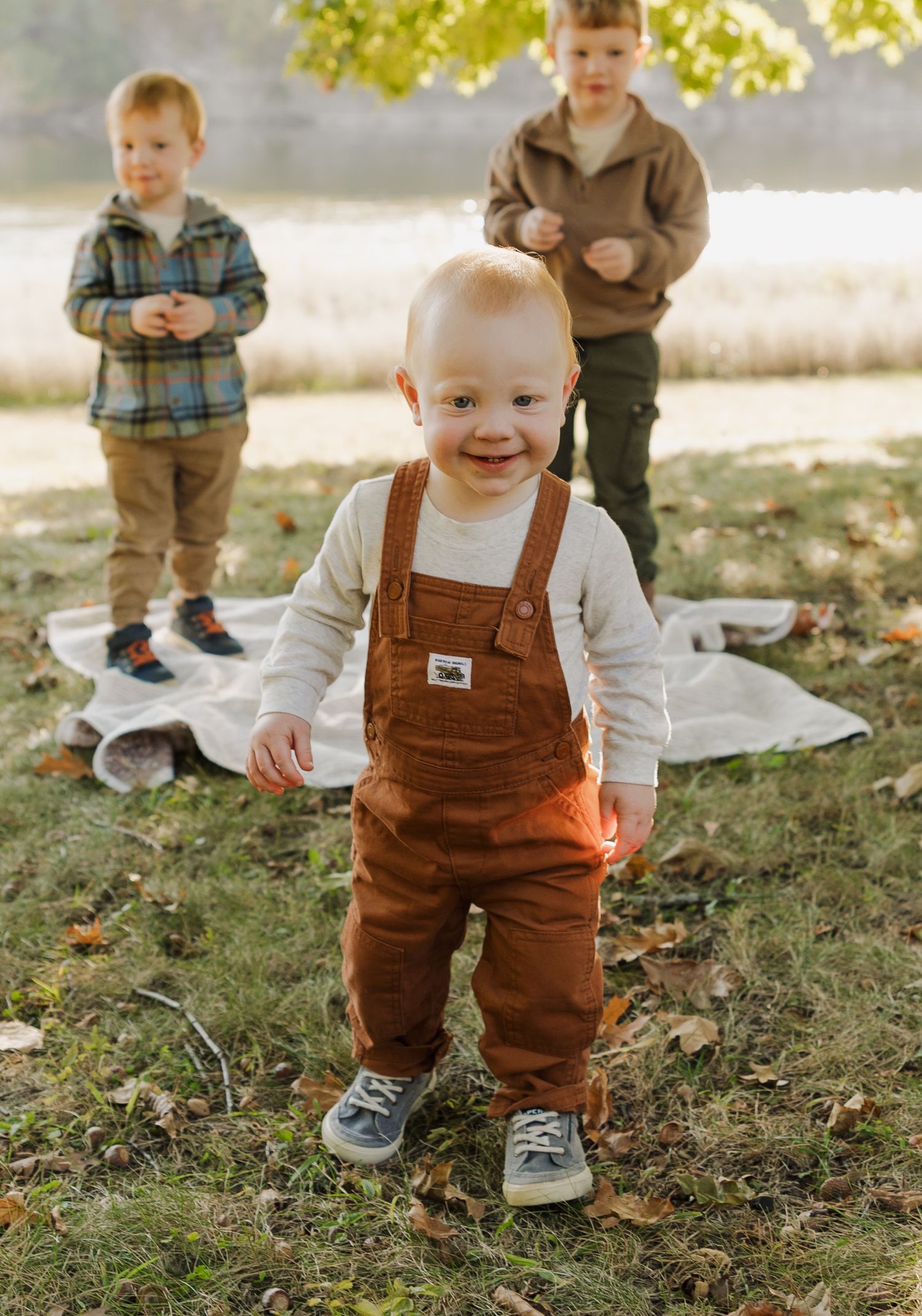 Three children stand outdoors on a blanket in autumn, with the front child wearing brown overalls and a white shirt.
