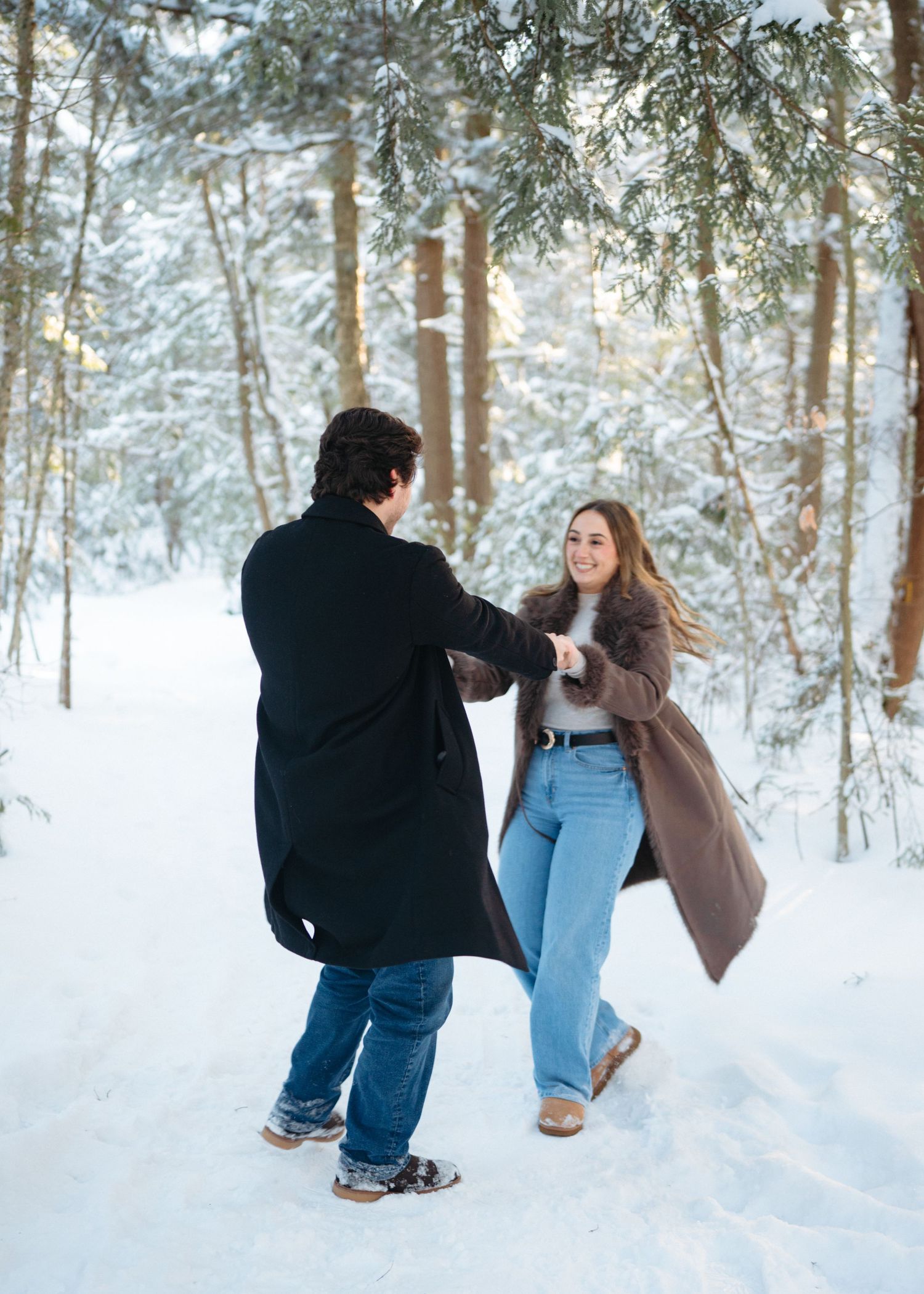 A couple dances playfully together in a snow-covered forest wearing winter coats and casual attire.