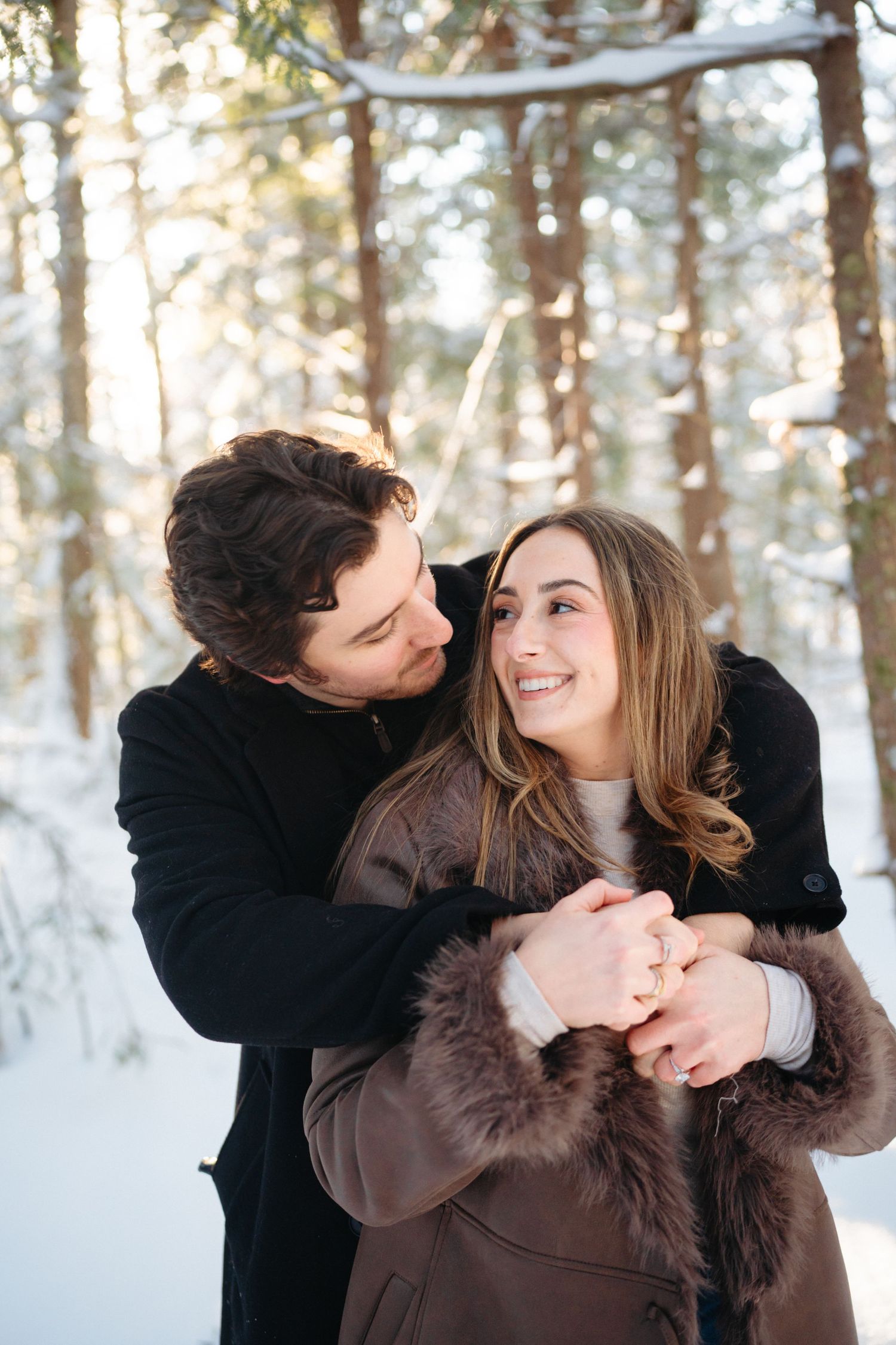 A couple embraces lovingly in a snowy winter forest setting with pine trees in the background.