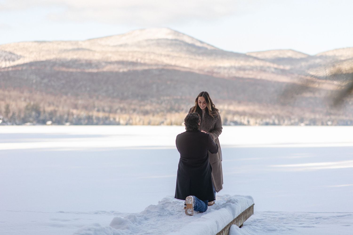 A snow-covered dock extends into a frozen lake with mountains visible in the distance.