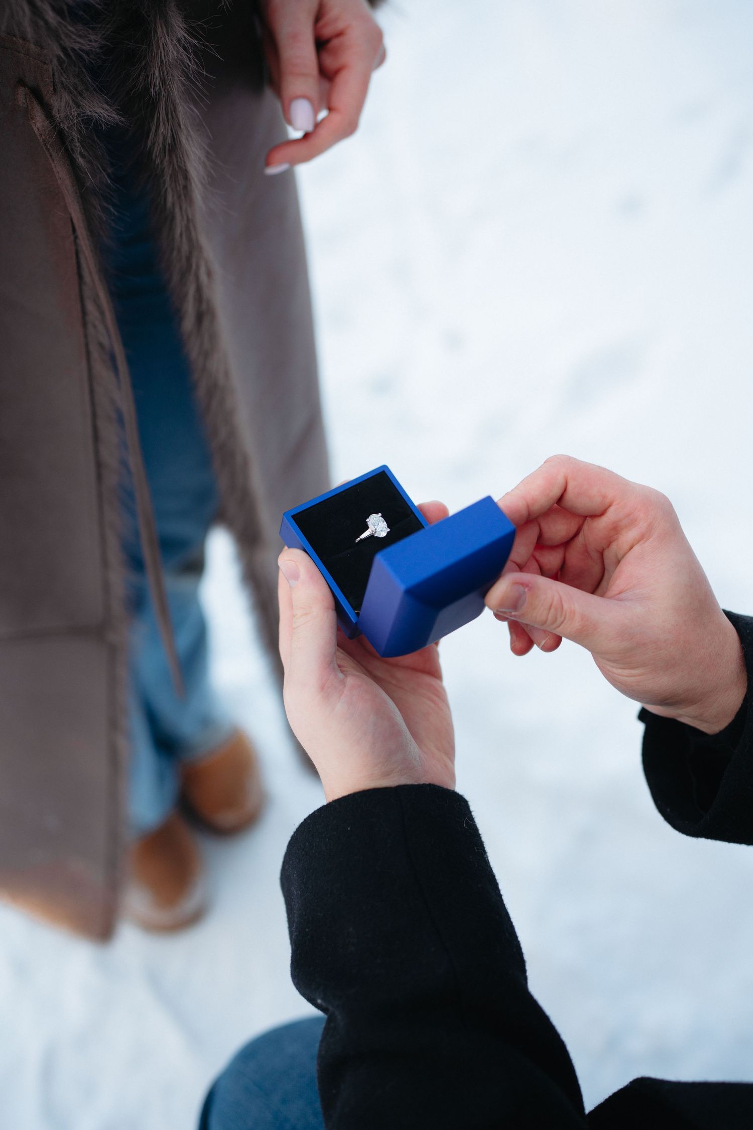 Hands holding a small blue ring box against a snowy white background during a proposal moment.