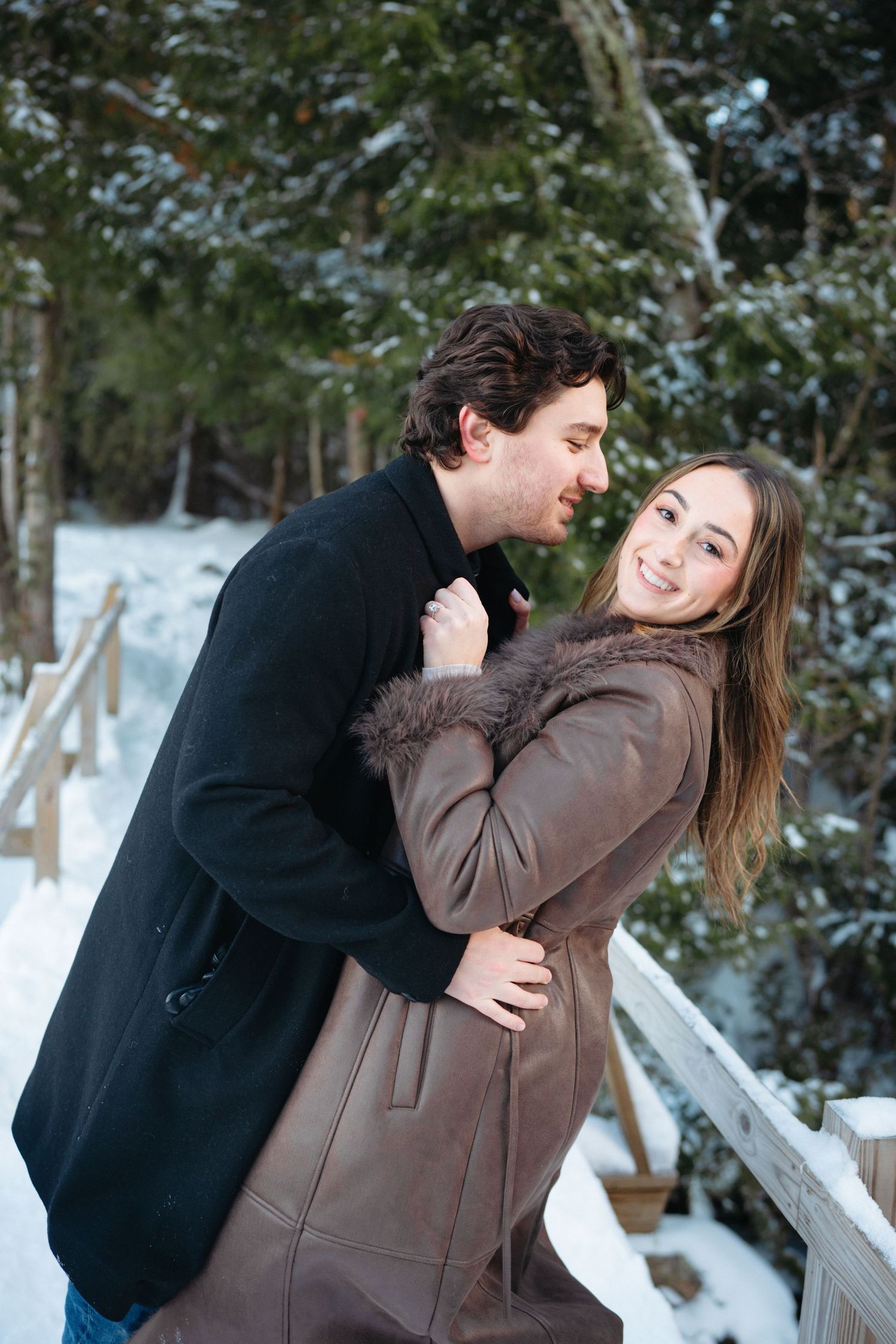 A couple embraces playfully in a snowy winter scene with evergreen trees in the background.