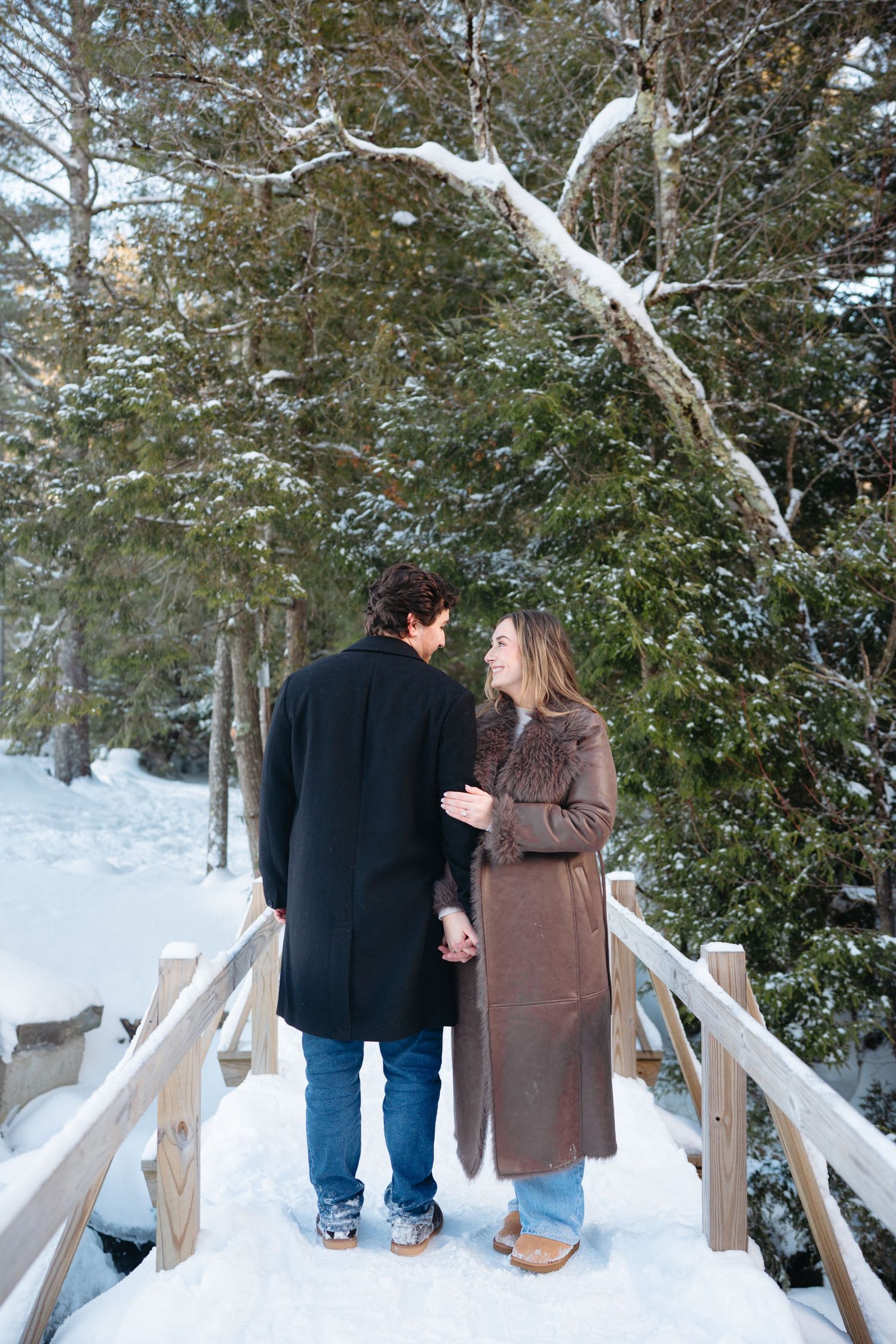 A couple shares a tender moment on a snow-covered wooden bridge in a winter forest.