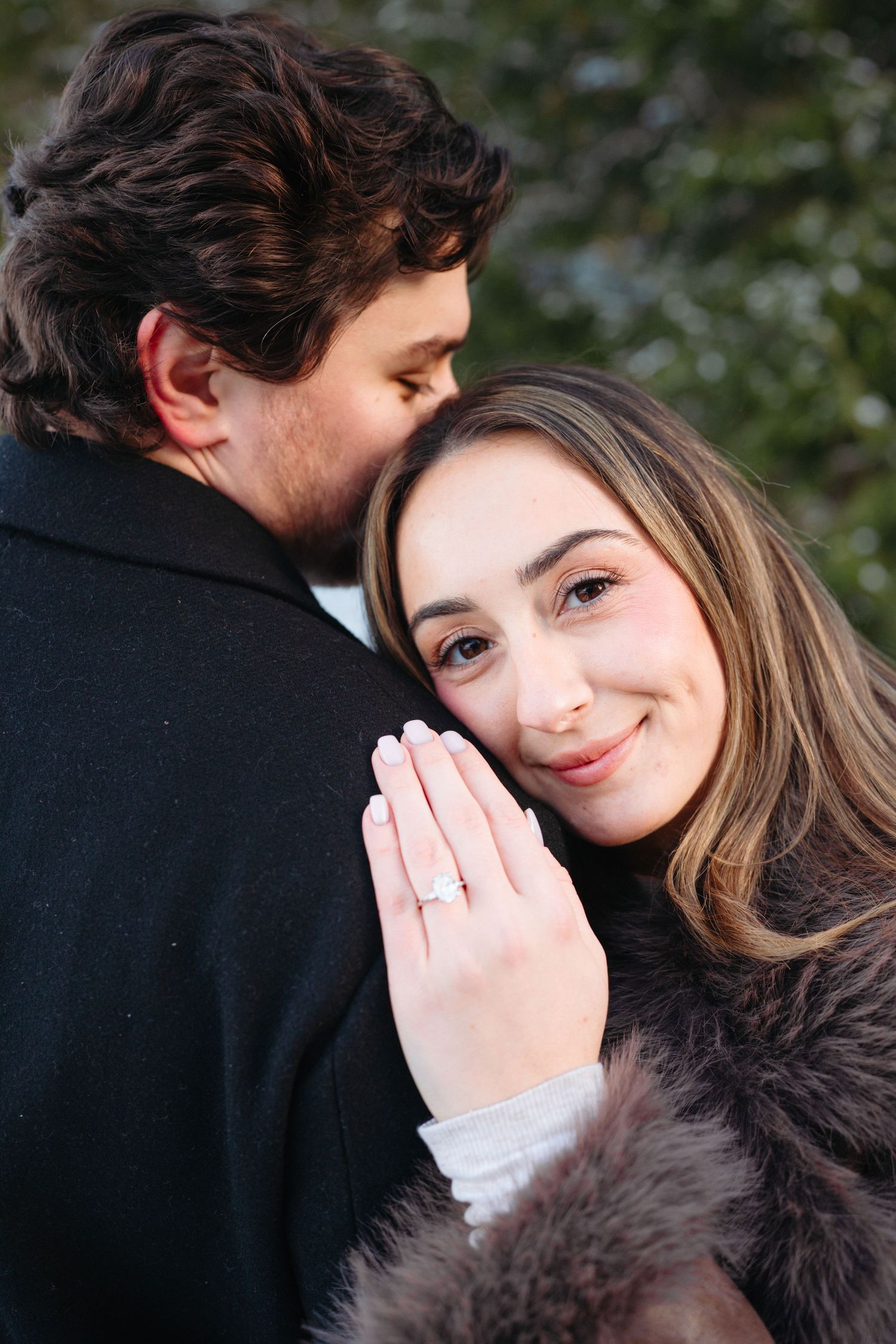 A newly engaged couple embraces while showing off an engagement ring in a romantic outdoor setting.