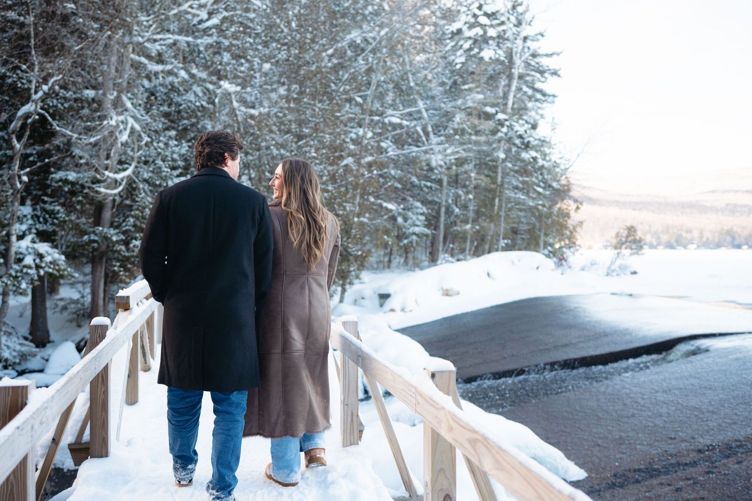 A couple walks together on a snowy bridge surrounded by frost-covered evergreen trees in winter.