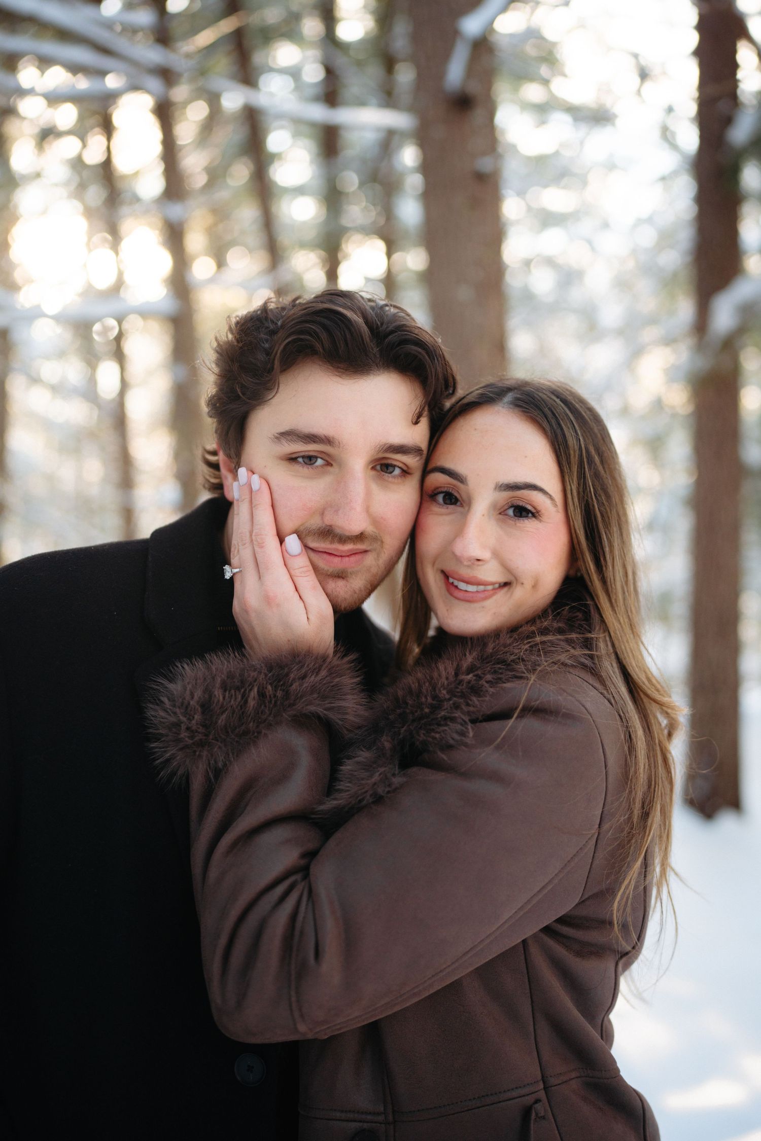 A couple in brown winter coats share affectionate moments in a snowy forest setting.
