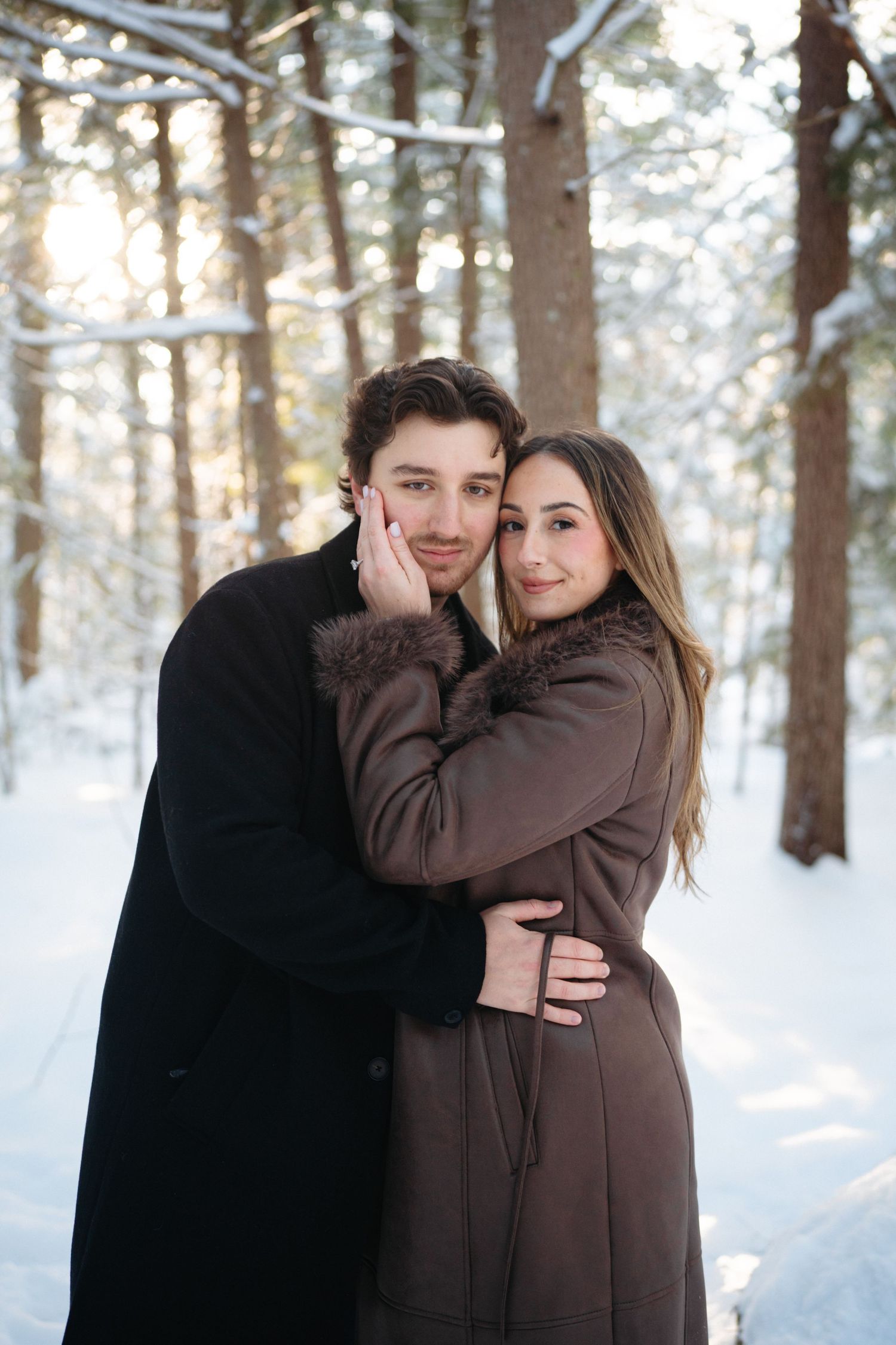 A couple in winter coats embraces while standing in a snowy forest with sunlight filtering through the trees.