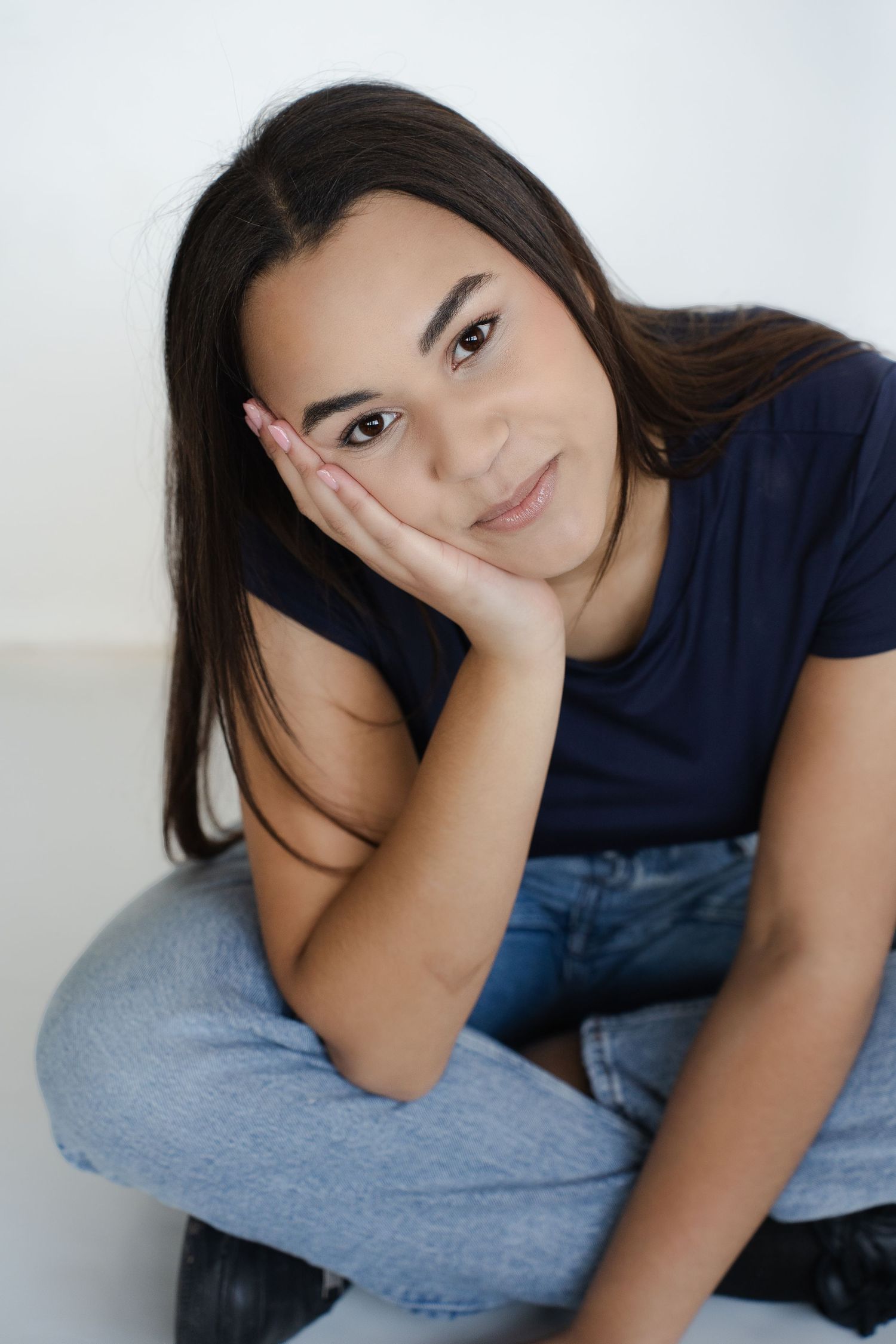 Young person in navy blue t-shirt and jeans sits casually against white wall with head resting on hand.