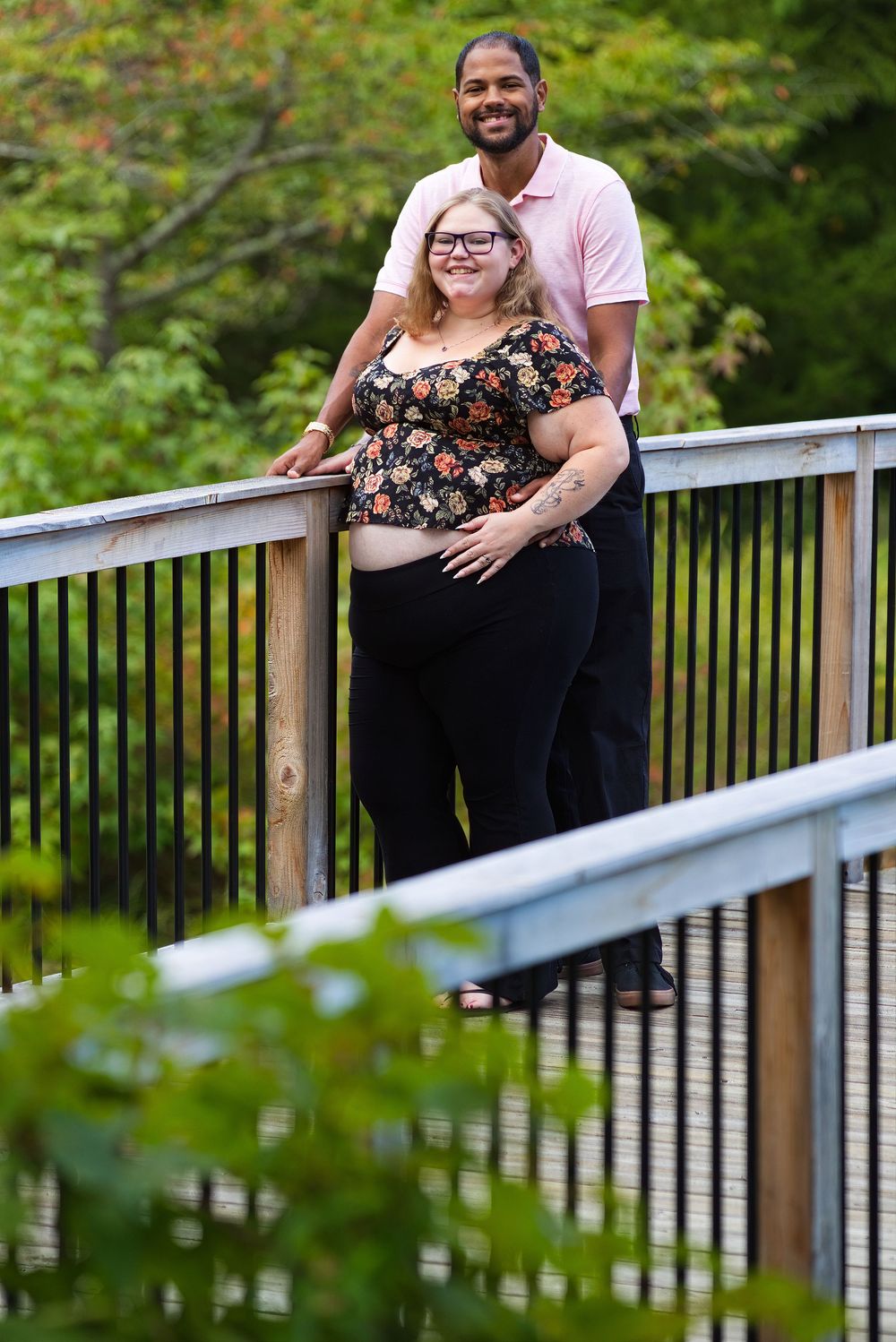 Couple posing on a wooden bridge surrounded by lush greenery, woman in floral top with baby bump.