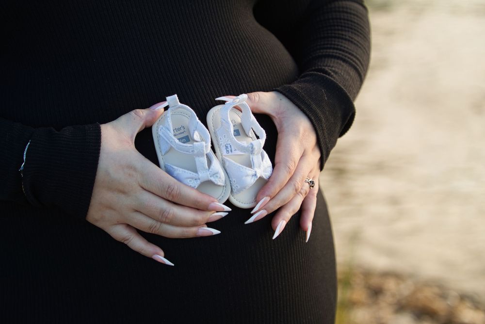 Pregnant woman in black dress holding tiny white baby shoes over her baby bump outdoors.