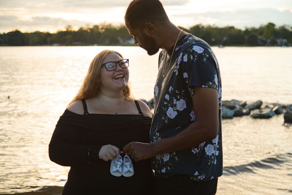 Happy couple holding tiny baby shoes by a lake at sunset, announcing their pregnancy outdoors.