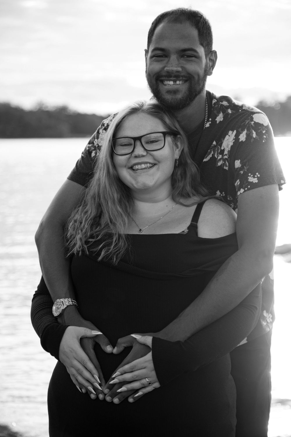 Black and white photo of a smiling couple embracing outdoors by a lake, making a heart shape with their hands.