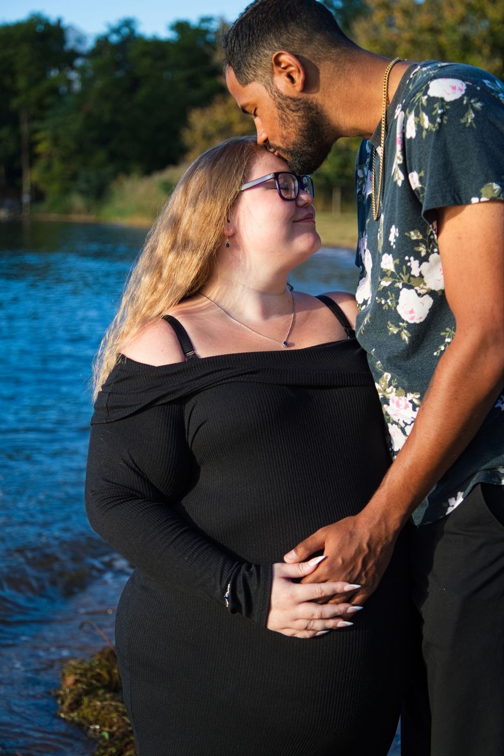 Pregnant couple sharing a tender moment by a lake, woman in black off-shoulder dress, man kissing her forehead.