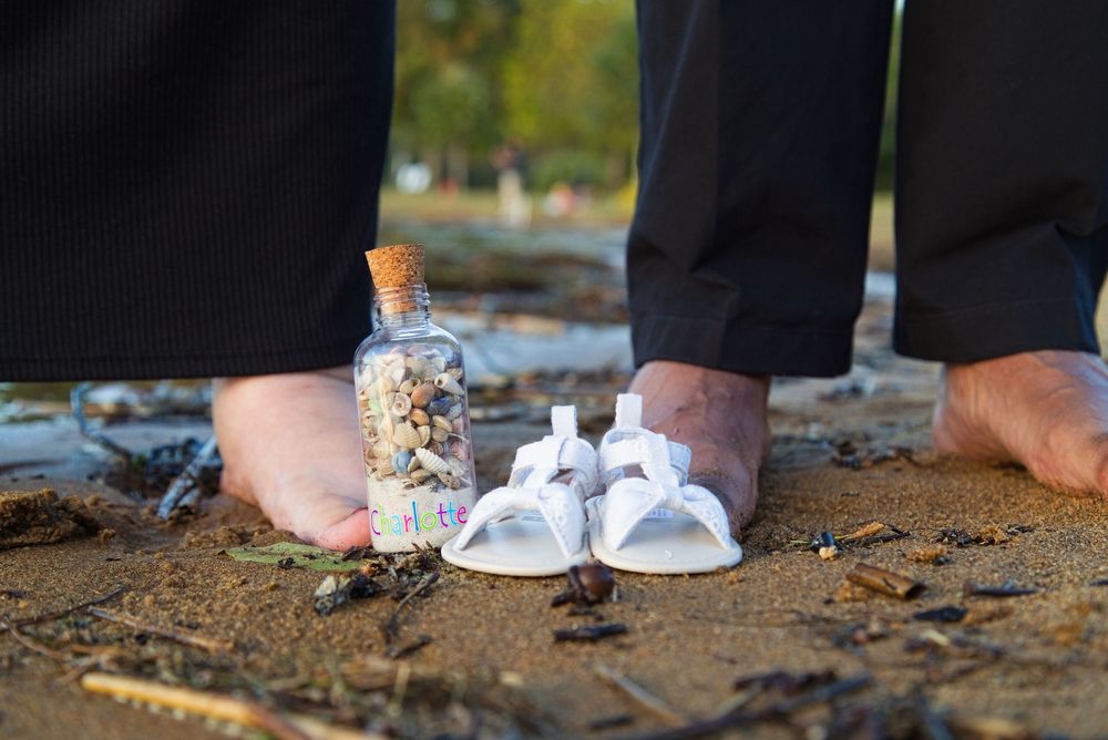 Bare adult feet beside tiny white baby shoes and a cork bottle filled with flowers on sandy ground outdoors.