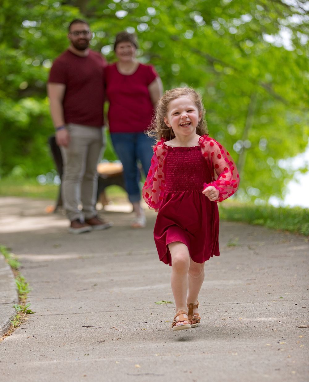 A joyful little girl in a red dress runs toward the camera while her parents smile in the background on a park path.