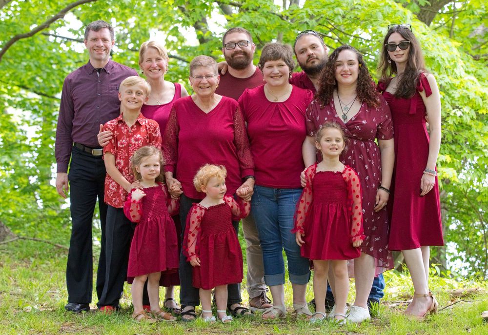 Large family group posing outdoors in matching red outfits, smiling together among green trees in a coordinated portrait.