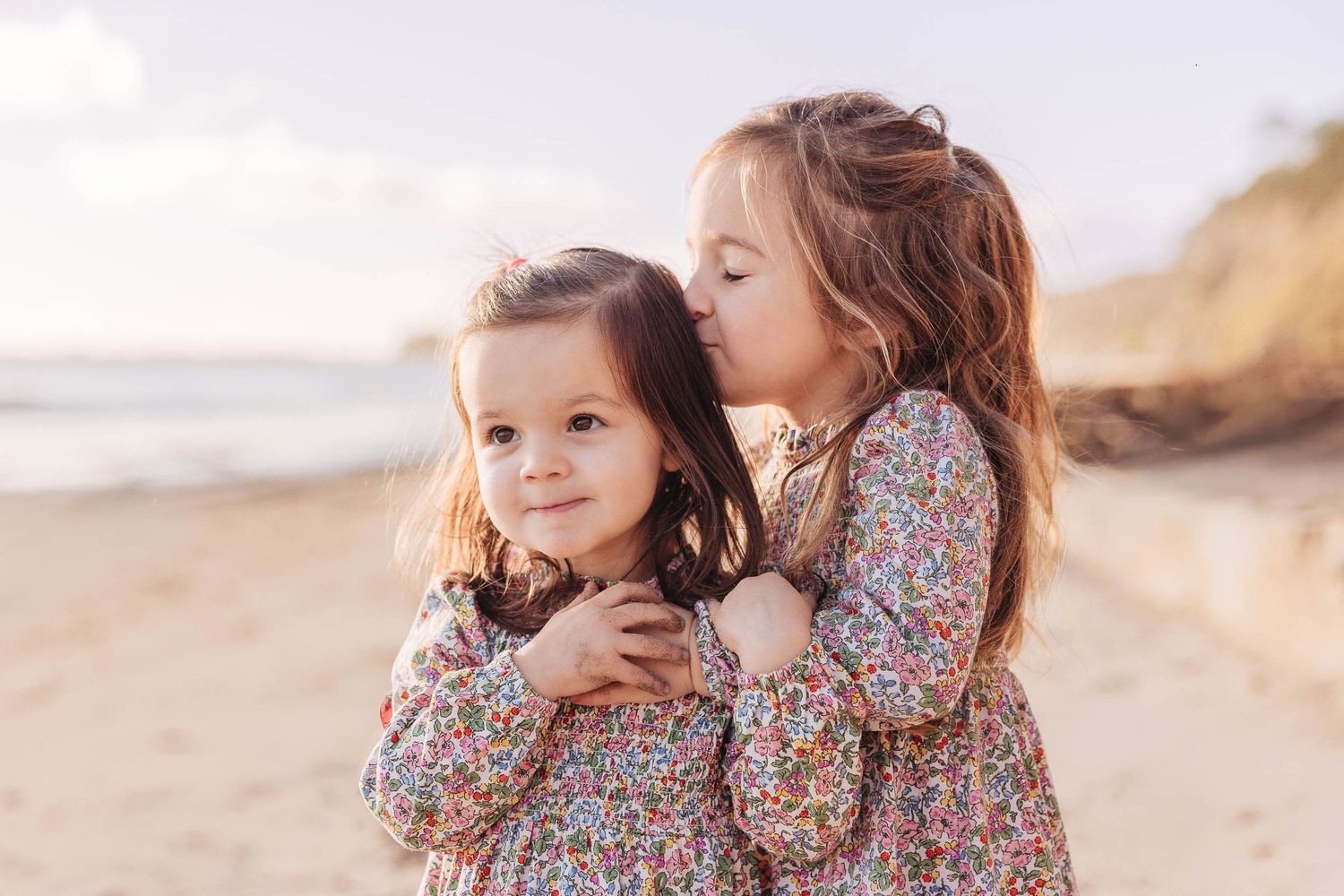 Mother and daughter in matching floral dresses share tender moments while walking along a sandy beach at sunset.