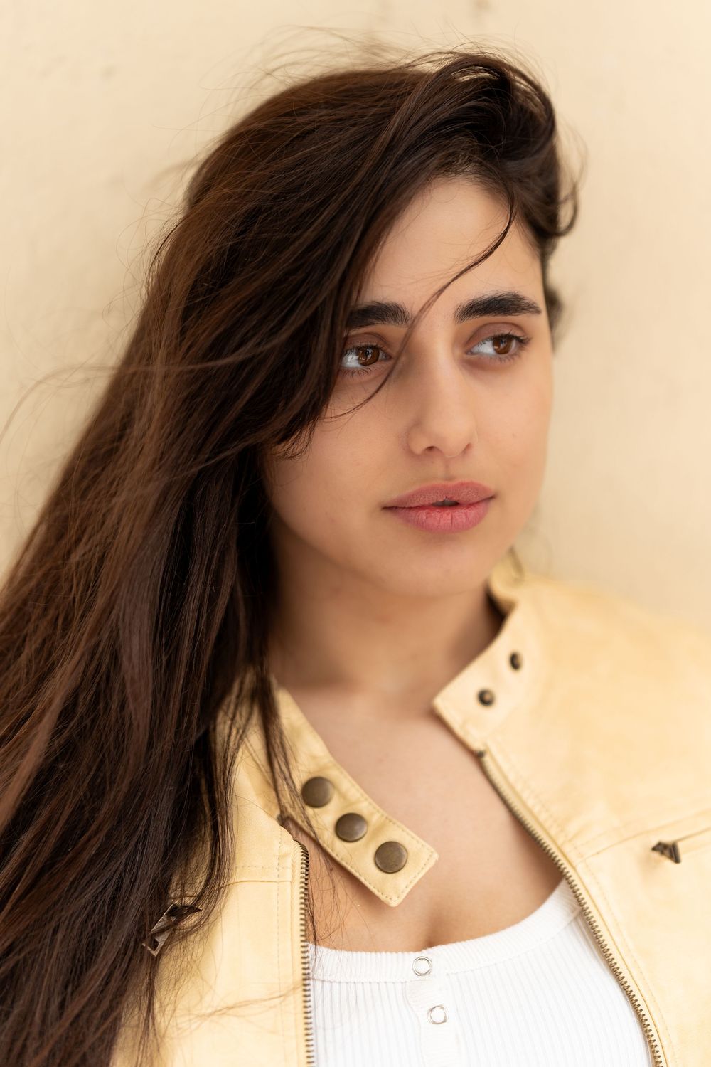 A fashion model in a cream leather jacket poses against a beige background with dark flowing hair and pink lipstick.