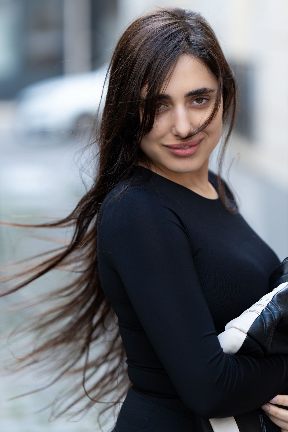 Person in black long sleeve top smiling and posing with windswept dark hair against blurred background.