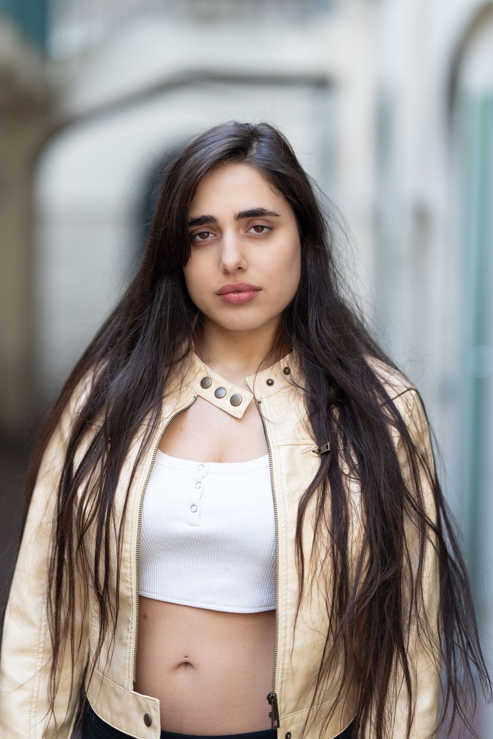 A person with long dark hair poses in a white crop top and beige jacket against a blurred background.