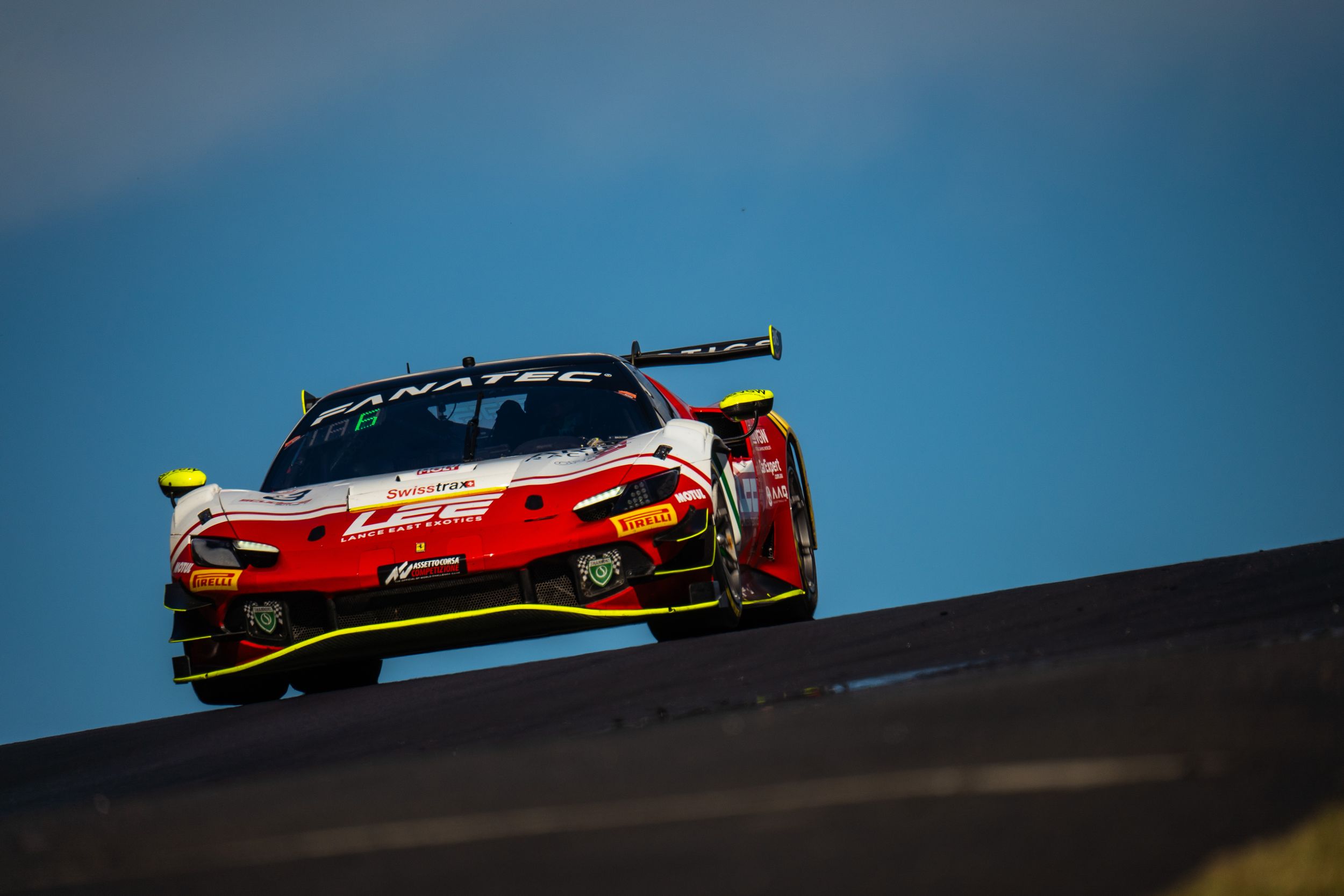 Red and black race cars compete on a racing track under clear blue skies during a professional motorsport event.
