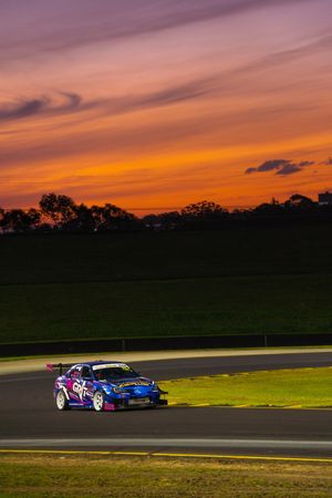 Race cars speed around a track against a vibrant orange and purple sunset sky with silhouetted trees in the background.