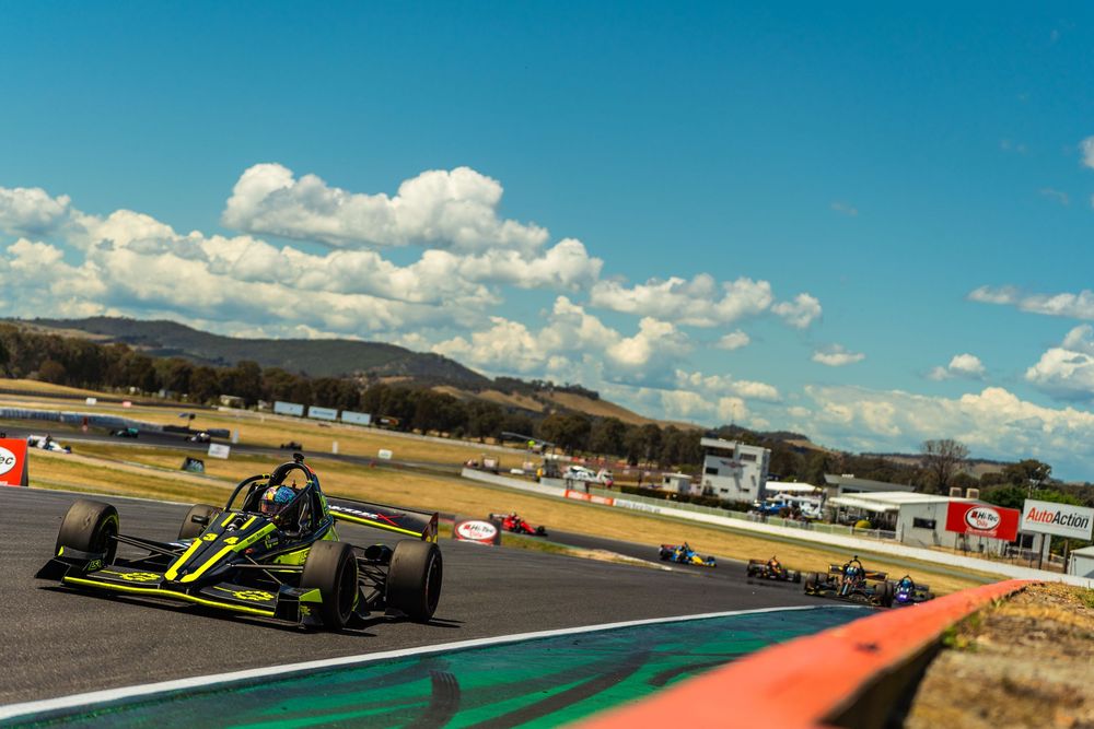 Race cars speed around a winding track during a motorsport competition under blue skies with white clouds.