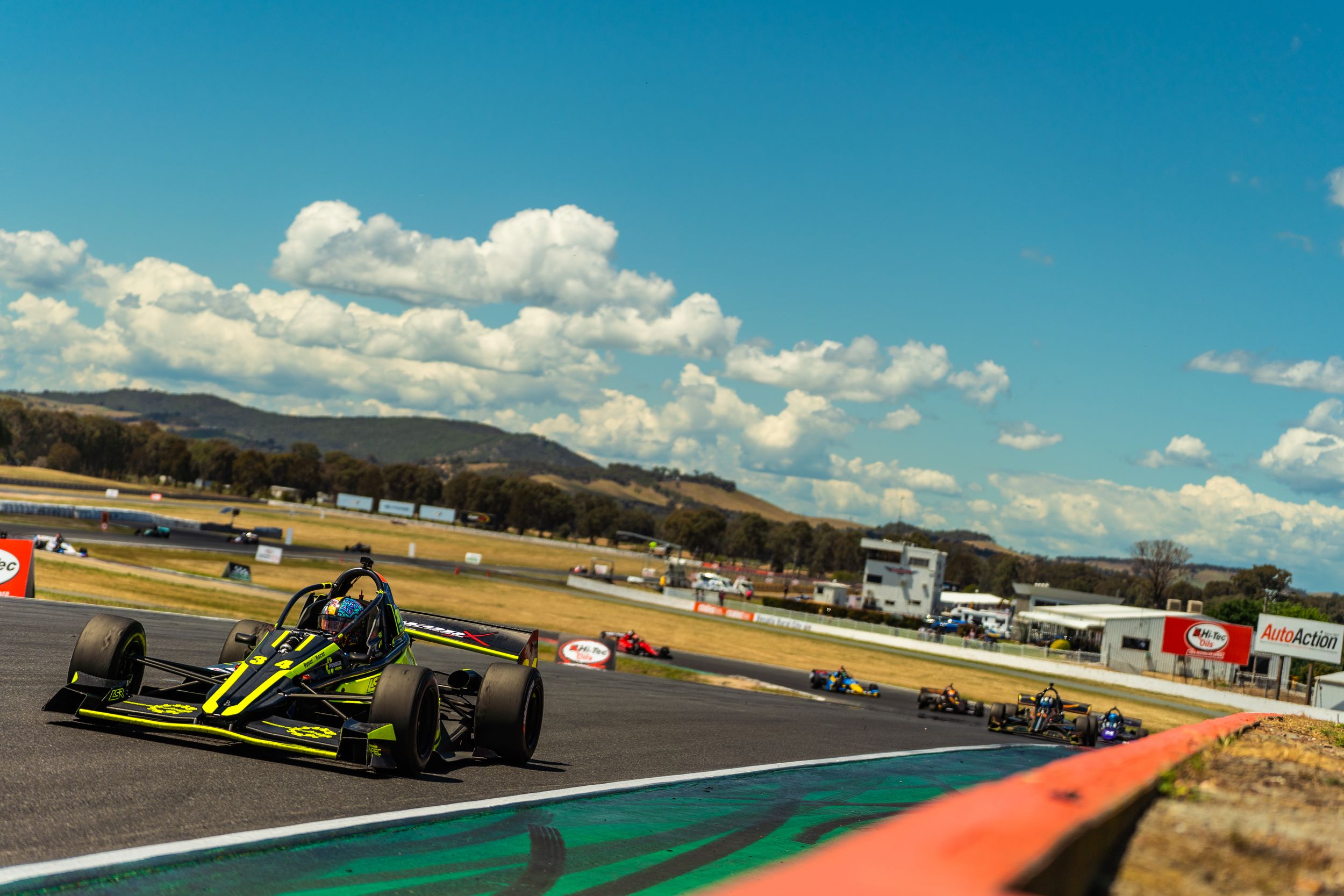 Race cars speed around a winding track during a motorsport competition under blue skies with white clouds.