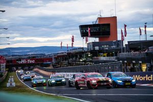 Race cars line up at the start of a motorsport event at a racing circuit with mountains visible in the background.