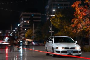 A white modified car drives on a wet urban street at night with autumn trees and city lights in the background.