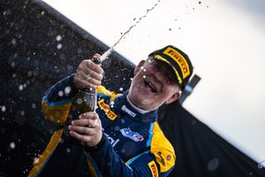 Racing driver celebrates by spraying champagne on the podium while wearing yellow team gear and racing suit.