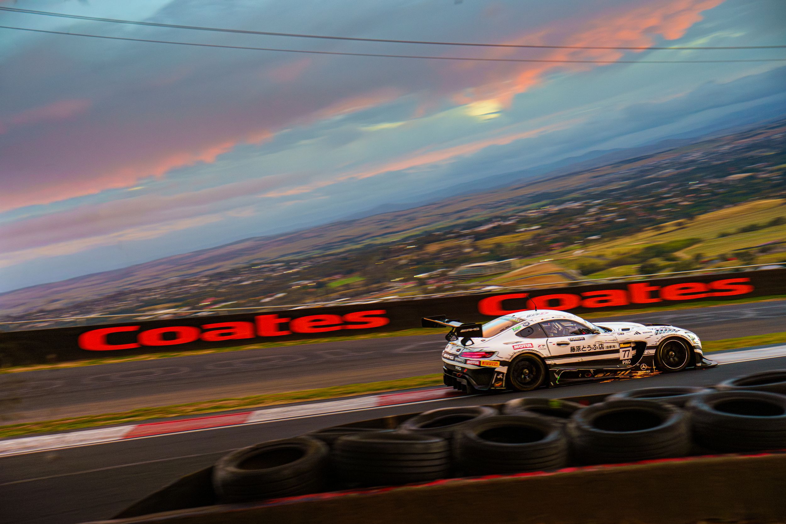 Race car speeds past tire barriers at sunset on Mount Panorama circuit with dramatic pink and blue sky overhead.