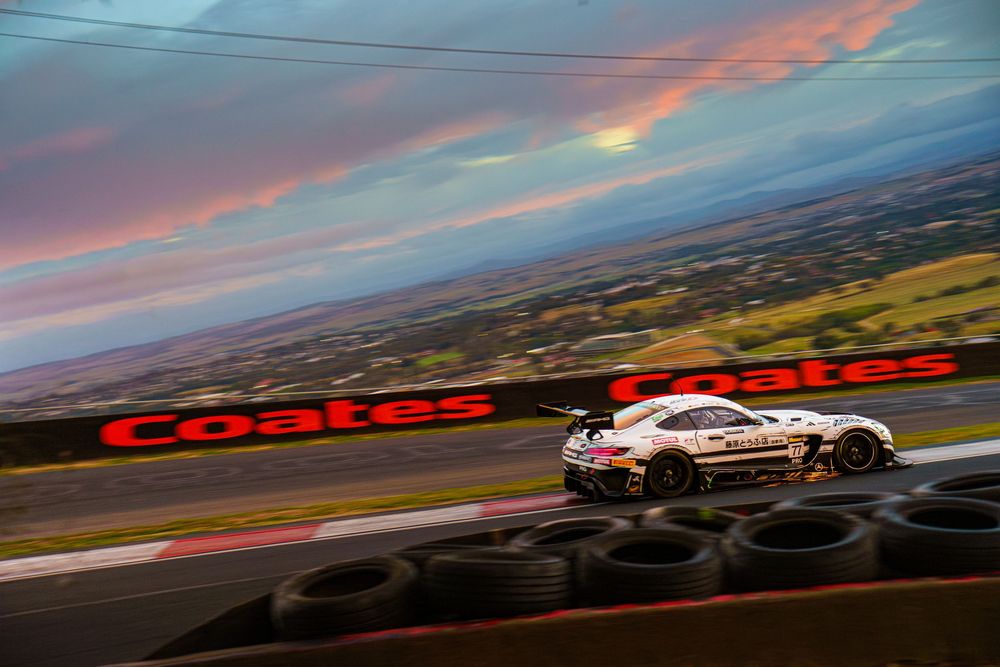 Race car speeds past tire barriers at sunset on Mount Panorama circuit with dramatic pink and blue sky overhead.
