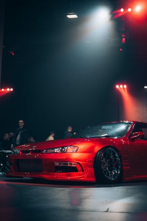 Modified red Nissan Silvia sports car with aftermarket wheels showcased under dramatic red and white stage lighting.