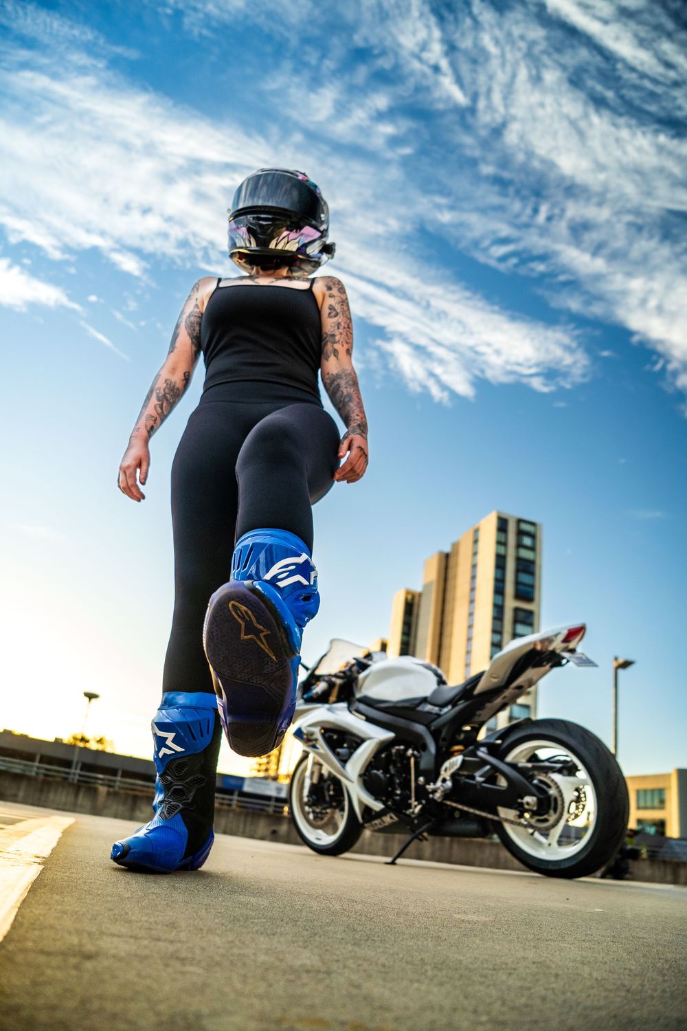 Motorcycle rider in black gear kneeling beside custom bike on urban rooftop with dramatic sky and buildings in background.