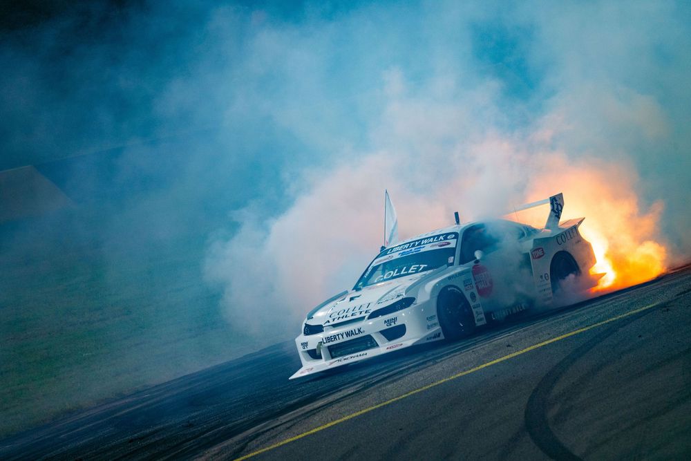 A race car drifts sideways on a track with smoke and flames coming from its exhaust during a dramatic motorsport event.
