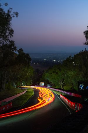 Long exposure photo of car light trails winding down Mount Panorama racing circuit at dusk with city lights below.