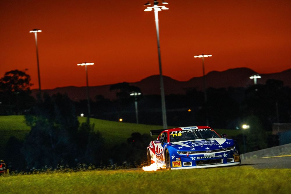 Race cars speed around an illuminated track at dusk with stadium lights glowing in the dark sky.