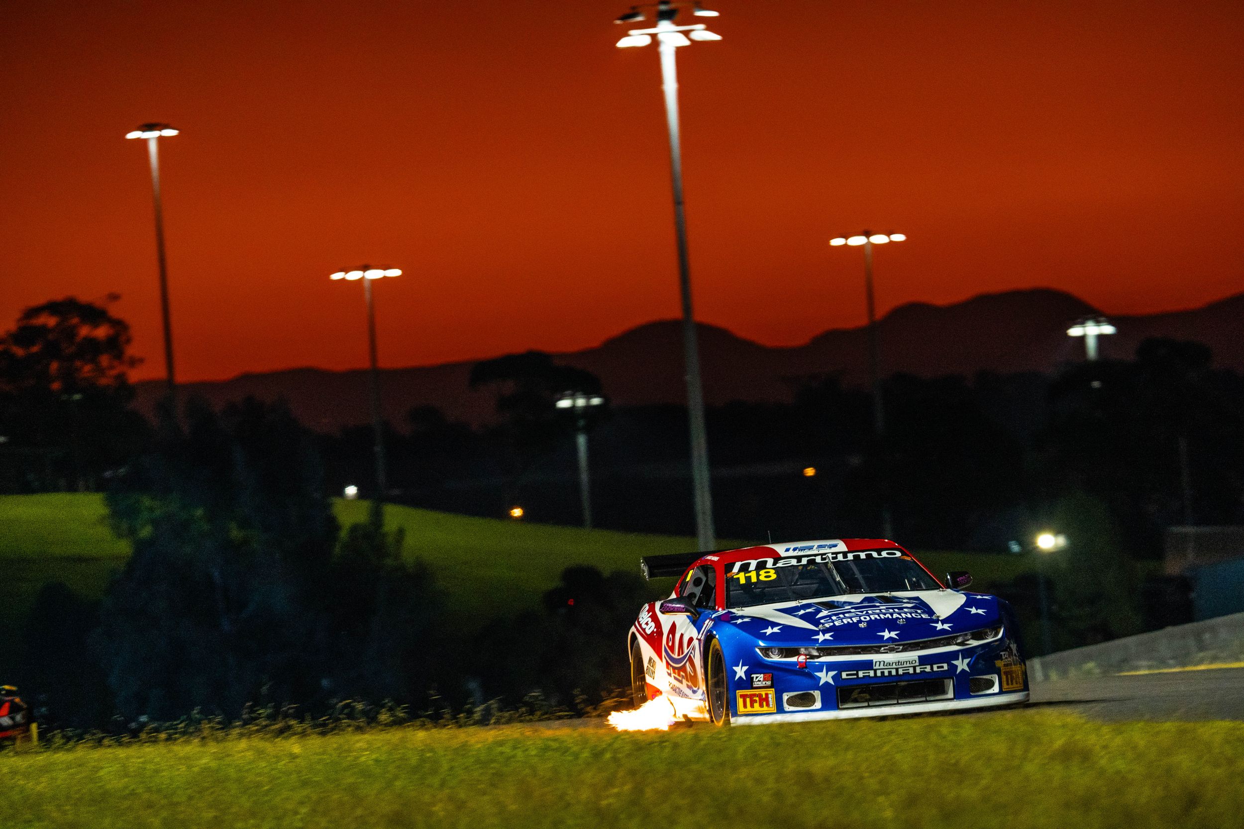 Race cars speed around an illuminated track at dusk with stadium lights glowing in the dark sky.
