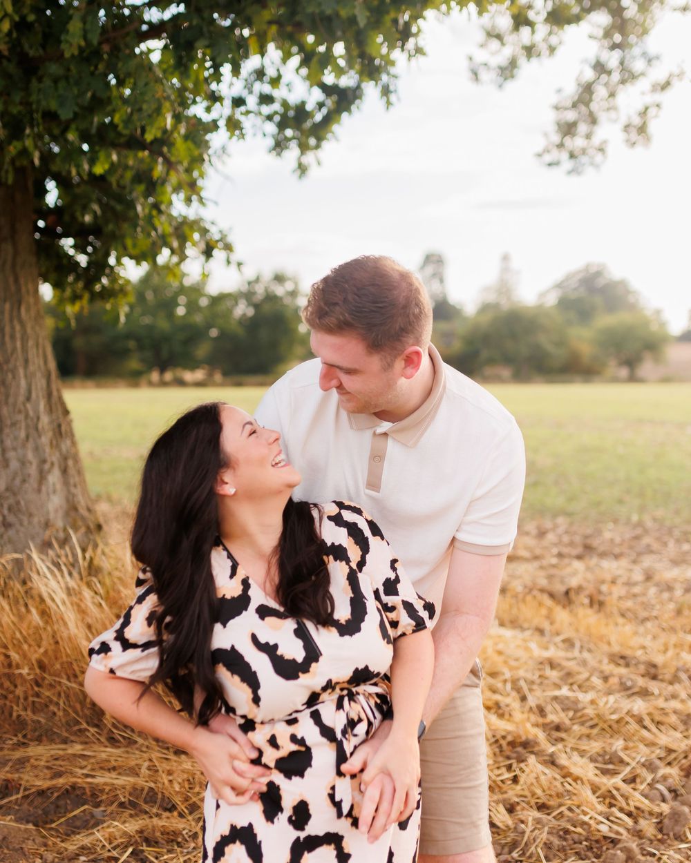A couple embraces and laughs together while sitting in a golden sunlit field during an outdoor photo session.